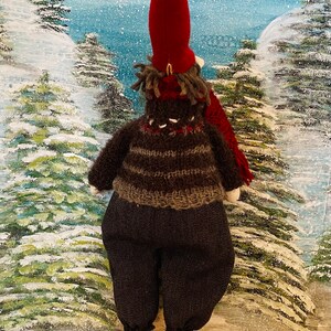 May include: A festive Christmas gnome ornament, featuring a long red hat and a brown and grey striped sweater. The gnome wears dark grey trousers, red and white striped legs, and red shoes. The background depicts a snowy winter scene.