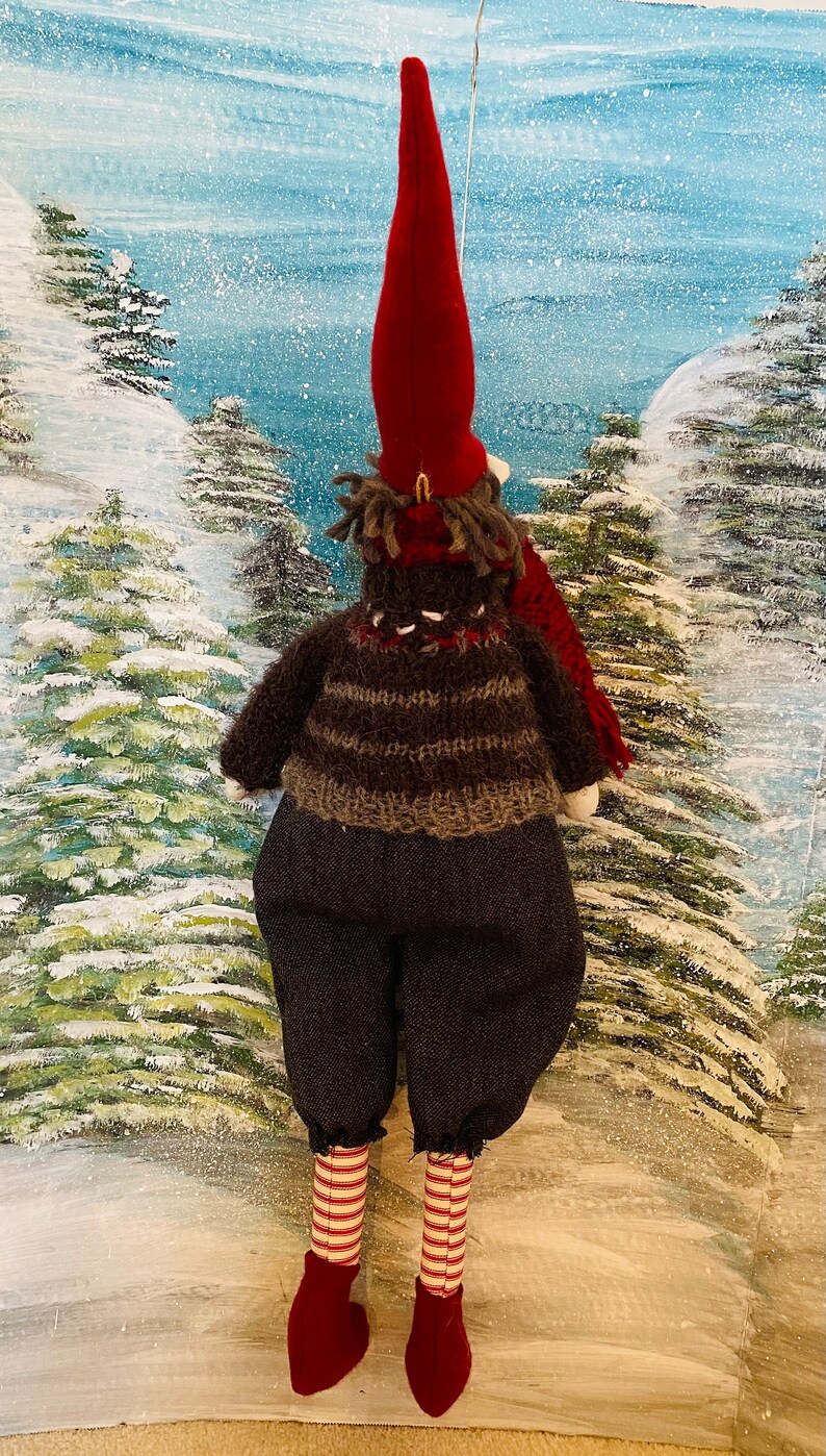 May include: A festive Christmas gnome ornament, featuring a long red hat and a brown and grey striped sweater. The gnome wears dark grey trousers, red and white striped legs, and red shoes. The background depicts a snowy winter scene.