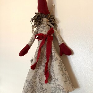 以下が含まれることがあります： 手作りの布製人形。長いとがったバーガンディ色の帽子と、お揃いのミトンを着用。人形のドレスはグレーの花柄で、バーガンディ色のリボンで結ばれています。人形はカールした茶色の髪をしています。