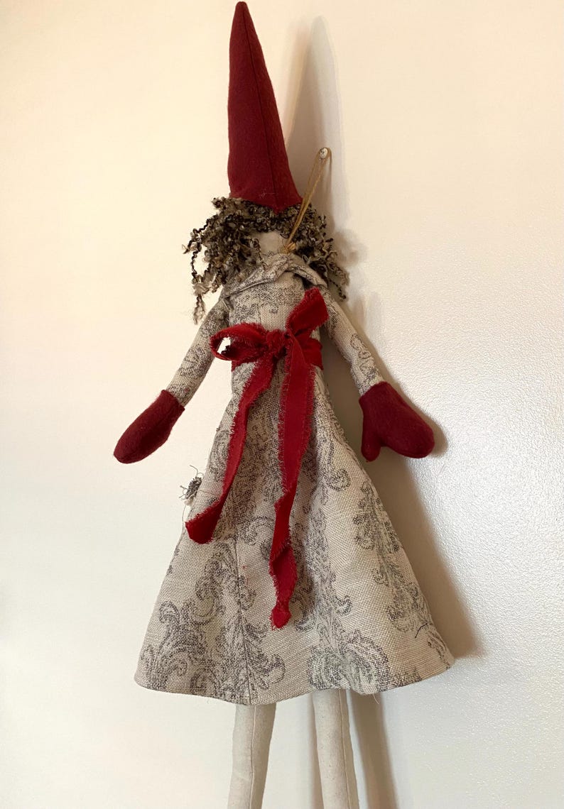 以下が含まれることがあります： 手作りの布製人形。長いとがったバーガンディ色の帽子と、お揃いのミトンを着用。人形のドレスはグレーの花柄で、バーガンディ色のリボンで結ばれています。人形はカールした茶色の髪をしています。