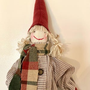 May include: A handcrafted doll featuring a red pointed hat, white yarn hair, and a smiling face. The doll is adorned with a plaid scarf in red, green, and beige, and a striped shirt with two brown buttons. The doll's arms and hat are red.