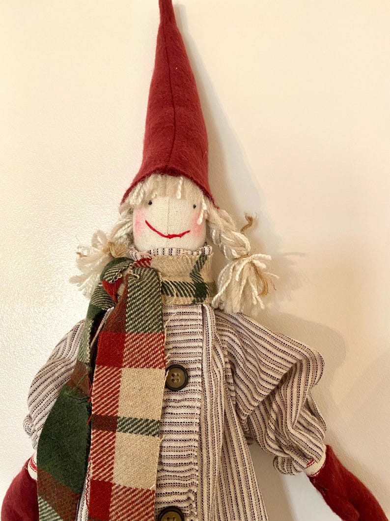 May include: A handcrafted doll featuring a red pointed hat, white yarn hair, and a smiling face. The doll is adorned with a plaid scarf in red, green, and beige, and a striped shirt with two brown buttons. The doll's arms and hat are red.