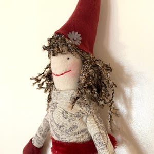 以下が含まれることがあります： 赤い尖った帽子と巻き毛の茶色い髪の毛を持つ手作りの人形。人形は、赤いベルベットのサッシュとマッチするミトンが付いた模様のドレスを着ています。小さな灰色の花が帽子を飾っています。
