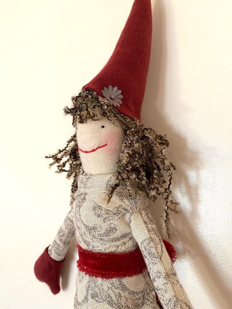 以下が含まれることがあります： 赤い尖った帽子と巻き毛の茶色い髪の毛を持つ手作りの人形。人形は、赤いベルベットのサッシュとマッチするミトンが付いた模様のドレスを着ています。小さな灰色の花が帽子を飾っています。