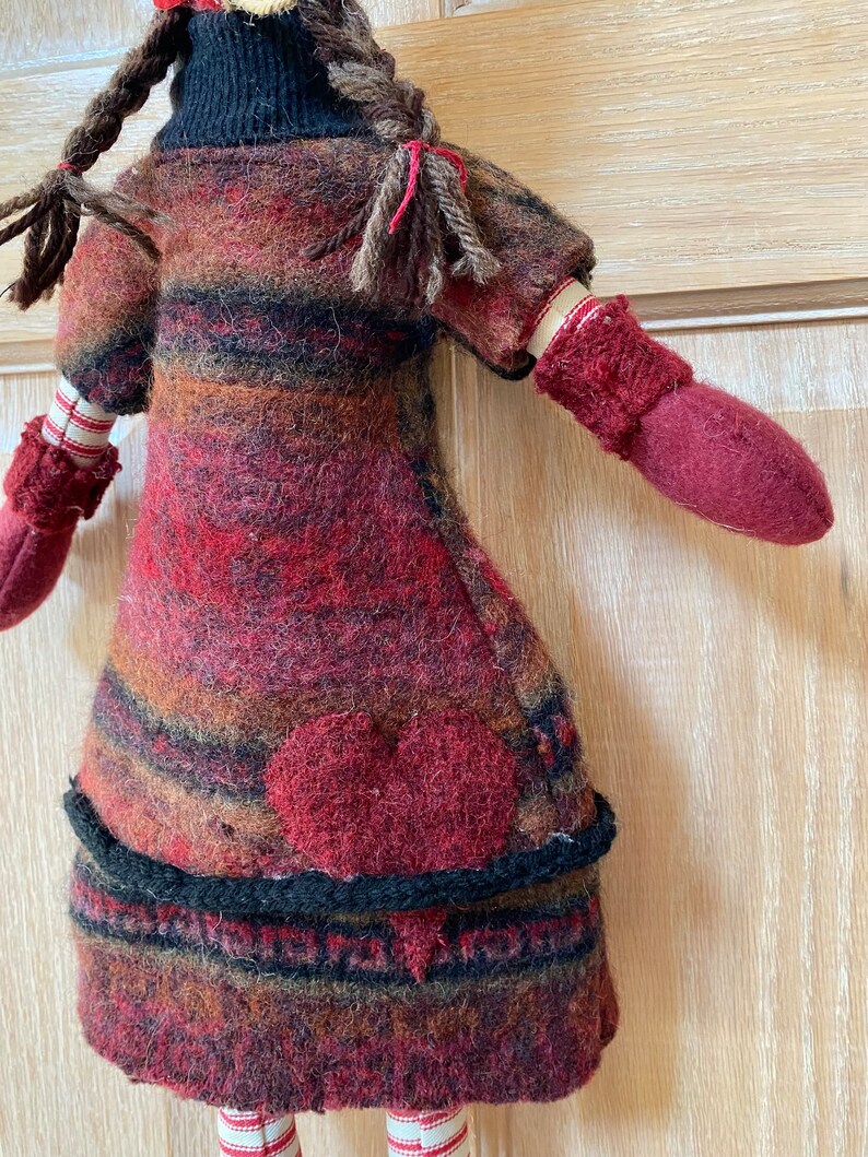 Peut inclure: Poup&eacute;e faite &agrave; la main avec une application de c&oelig;ur rouge sur le devant. La poup&eacute;e porte une robe ray&eacute;e dans les tons de rouge, marron et noir, avec des mitaines assorties. La poup&eacute;e a des cheveux bruns tress&eacute;s et des jambes ray&eacute;es.