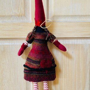 Peut inclure: Poup&eacute;e de No&euml;l faite &agrave; la main, avec un grand chapeau pointu bordeaux et des mitaines et chaussures assorties. La robe de la poup&eacute;e est un m&eacute;lange de laine bordeaux, marron et noire. Ses jambes sont ray&eacute;es rouge et blanc. D&eacute;coration de vacances festive.