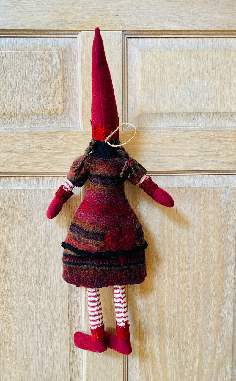 Peut inclure: Poup&eacute;e de No&euml;l faite &agrave; la main, avec un grand chapeau pointu bordeaux et des mitaines et chaussures assorties. La robe de la poup&eacute;e est un m&eacute;lange de laine bordeaux, marron et noire. Ses jambes sont ray&eacute;es rouge et blanc. D&eacute;coration de vacances festive.