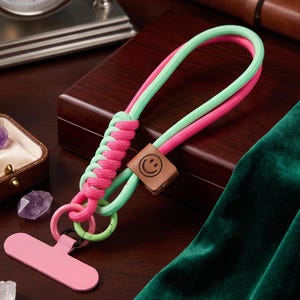 May include: A phone lanyard featuring intertwined mint green and hot pink paracord, a square brown leather charm embossed with a smiley face, and a solid pink rectangular phone tether tab attached to a circular ring at the base of the cord.