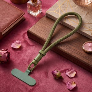 May include: An olive green phone tether tab with a matching braided cord wrist strap. The strap features a sturdy olive green plastic swivel clasp that connects the cord to the flat, T-shaped adhesive tab designed for attachment to a mobile device.