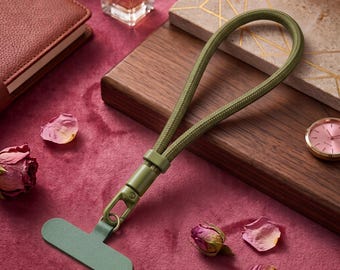 May include: An olive green phone tether tab with a matching braided cord wrist strap. The strap features a sturdy olive green plastic swivel clasp that connects the cord to the flat, T-shaped adhesive tab designed for attachment to a mobile device.