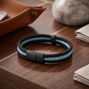 Könnte beinhalten: Ein geflochtenes Armband aus schwarzen und hellblauen Kordeln mit einem mattschwarzen Metallverschluss. Das Armband besteht aus zwei dicken schwarzen Außenbändern und einem hellblauen Mittelband, die durch einen rechteckigen schwarzen Verschluss verbunden sind.