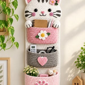 Könnte beinhalten: Ein weißer, gehäkelter Wand-Organizer in Katzenform mit drei Taschen. Die obere Tasche ist rosa mit einer weißen Blume, die mittlere ist grau mit einem rosa Herz und die untere ist rosa mit einem rosa Pfotenabdruck. Er hängt an einem Holzhaken.