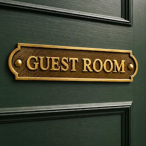May include: A rectangular brass plaque with rounded ends and a textured dark bronze center, featuring the raised gold-colored text GUEST ROOM. The sign is secured with two visible brass screws on a dark green paneled door surface.