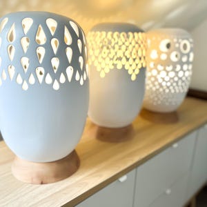 Może przedstawiać: Trzy ceramiczne lampy z białym szkliwem i misternymi wzorami wyciętymi. Lampy stoją na drewnianej powierzchni i mają ciepłe światło.