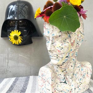 May include: A white ceramic bust vase with colorful paint splatters, filled with a variety of flowers, including yellow, orange, and purple. A black Darth Vader mask is on the left side of the image.