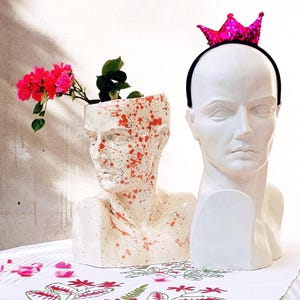 May include: Two white plaster busts, one with orange and white paint splatters, used as planters. The bust with splatters has a small plant growing out of the top. The other bust has a pink sparkly crown headband on top. Both busts are sitting on a white tablecloth with pink flowers.