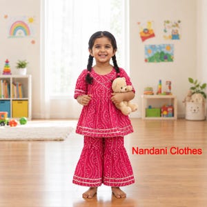 May include: A bright pink two-piece outfit featuring a short-sleeved peplum top and flared pants with a white dotted pattern and metallic trim. The outfit is paired with a light brown teddy bear. The text Nandani Clothes appears in red.
