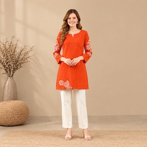 May include: A bright orange tunic featuring three-quarter length sleeves with white floral embroidery and a small notch neckline, paired with straight-leg white trousers. The tunic has a subtle white dotted pattern across the front fabric.