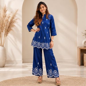 May include: A royal blue two-piece set consisting of a tunic and wide-leg trousers. The tunic features a mandarin collar, three-quarter sleeves, and white floral embroidery. The hem of the tunic and the trousers have a scalloped white embroidered edge.