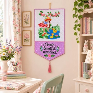 May include: A quilted fabric wall hanging featuring an embroidered illustration of a child in a red polka dot dress near a pond with yellow ducks. The lower purple section displays the embroidered text Create beautiful memories with floral accents.
