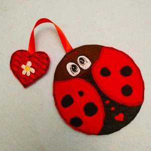 Puede incluir: Una mariquita roja con manchas negras y una cabeza marrón con ojos blancos. La mariquita está unida a una cinta roja con un corazón rojo con una flor blanca.