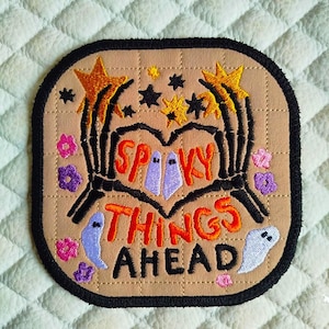May include: Embroidered patch with a tan background and a black border. The patch features the words "Spooky Things Ahead" in orange, with skeleton hands forming a heart shape. Includes stars, flowers, and ghost designs in various colors.