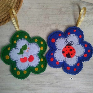 May include: Two felt flower-shaped ornaments with embroidered designs. One ornament is green with yellow polka dots and features a cherry design. The other ornament is blue with red polka dots and features a ladybug design.
