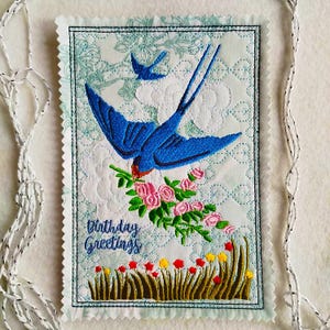 May include: Embroidered birthday card with a blue swallow carrying pink roses. The card has a light blue floral background and the words "Birthday Greetings" are written in cursive. Yellow and red flowers are at the bottom.