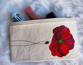 May include: A cream-coloured zipped pouch featuring a large embroidered red poppy flower design. The pouch contains a pink makeup brush, a multi-colour pen, and a black lipstick tube. The pouch is on a white, fluffy surface.
