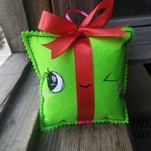 May include: A green felt gift box with a red ribbon and bow. The box has a winking cartoon face with eyelashes and a smile.