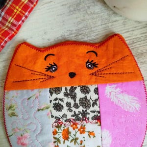 Pu&ograve; includere: Un sottobicchiere rotondo in tessuto a forma di gatto arancione con un design patchwork. Il viso del gatto &egrave; ricamato con baffi neri, un naso e occhi rosa.