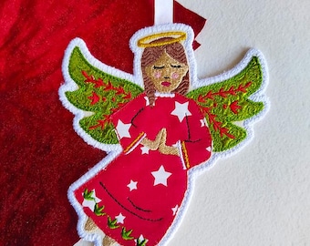 Puede incluir: Adorno de ángel bordado con un vestido rojo adornado con estrellas blancas y detalles verdes y rojos. El ángel tiene cabello castaño, un halo dorado y alas verdes con detalles rojos. Se adjunta una cinta blanca para colgar.
