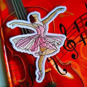 Può includere: Toppa ricamata raffigurante una ballerina in tutù rosa, con le braccia tese, con bordo bianco. La toppa è ambientata su uno sfondo rosso con un violino e note musicali, creando un tema di danza e musica.