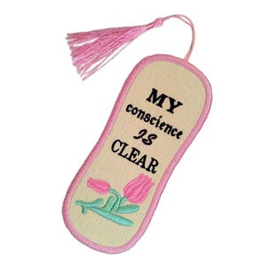 May include: Embroidered bookmark with a light beige background, pink trim, and a pink tassel. The bookmark features the text "MY conscience IS CLEAR" in black, along with an embroidered design of pink tulips with green stems and leaves.