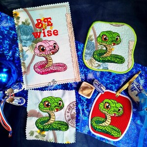 Könnte beinhalten: Vier gestickte Patches mit Cartoon-Schlangen. Ein Patch ist pink und sagt "Be Wise". Die anderen drei Patches sind grün und zeigen eine Schlange mit herausgestreckter Zunge. Die Patches befinden sich auf einem blauen Hintergrund.