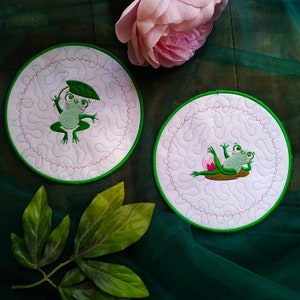 May include: Two round, white coasters with green embroidered edges. Each coaster features a green frog with a pink lily pad. One frog is sitting on the lily pad, while the other is holding a green leaf over its head.