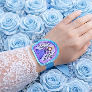 Puede incluir: Un parche bordado azul con un dise&ntilde;o de &aacute;ngel, usado en la mu&ntilde;eca con una cinta azul. El parche tiene detalles morados, azules y blancos. El fondo est&aacute; lleno de rosas azul claro.