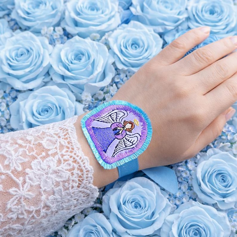 Puede incluir: Un parche bordado azul con un dise&ntilde;o de &aacute;ngel, usado en la mu&ntilde;eca con una cinta azul. El parche tiene detalles morados, azules y blancos. El fondo est&aacute; lleno de rosas azul claro.