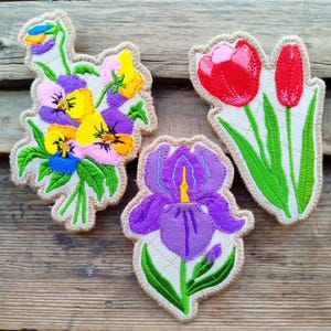 May include: Three embroidered floral patches: a bouquet of pansies in yellow, purple, and pink; a purple iris with a yellow center; and two red tulips with green stems and leaves. Each patch has a beige border and is set against a wooden background.