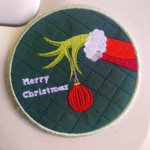 May include: A green fabric circle with a white stitched border. The center of the circle features an embroidered image of a green hand holding a red Christmas ornament. The text "Merry Christmas" is embroidered in purple below the ornament.