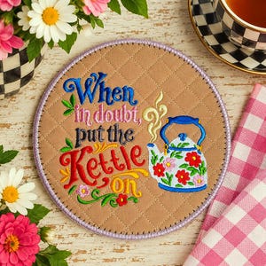 Könnte beinhalten: Ein runder, gesteppter beigefarbener Untersetzer mit gesticktem Text "When in doubt, put the Kettle on" in Blau, Pink, Schwarz und Rot. Rechts ist ein hellblauer Kessel mit Blumenmuster und gelbem Dampf zu sehen, eingefasst in Lila.