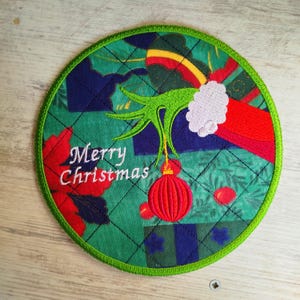 Puede incluir: Mano verde sosteniendo un adorno navideño rojo con el texto "Merry Christmas" sobre un fondo de tela acolchada verde, azul y roja.