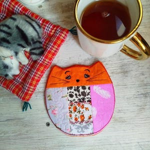 May include: A colorful patchwork cat-shaped coaster with an orange face and embroidered whiskers. The coaster is made of different fabrics and has a pink, purple, and white design.