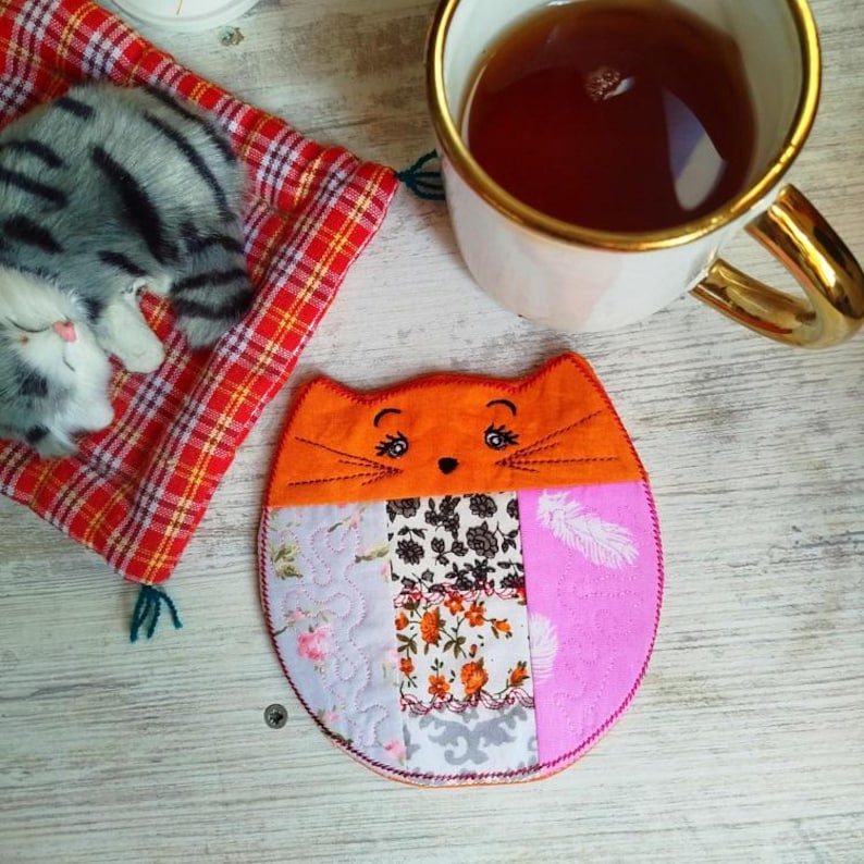 Pu&ograve; includere: Un sottobicchiere a forma di gatto fatto di patchwork colorato con un viso arancione e baffi ricamati. Il sottobicchiere &egrave; fatto di diversi tessuti e ha un design rosa, viola e bianco.