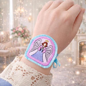 Puede incluir: Brazalete bordado con un dise&ntilde;o de &aacute;ngel. El brazalete tiene un borde azul claro y un interior rosa con un &aacute;ngel con un vestido azul, halo dorado y detalles rosas. Se asegura con cintas azul claro.