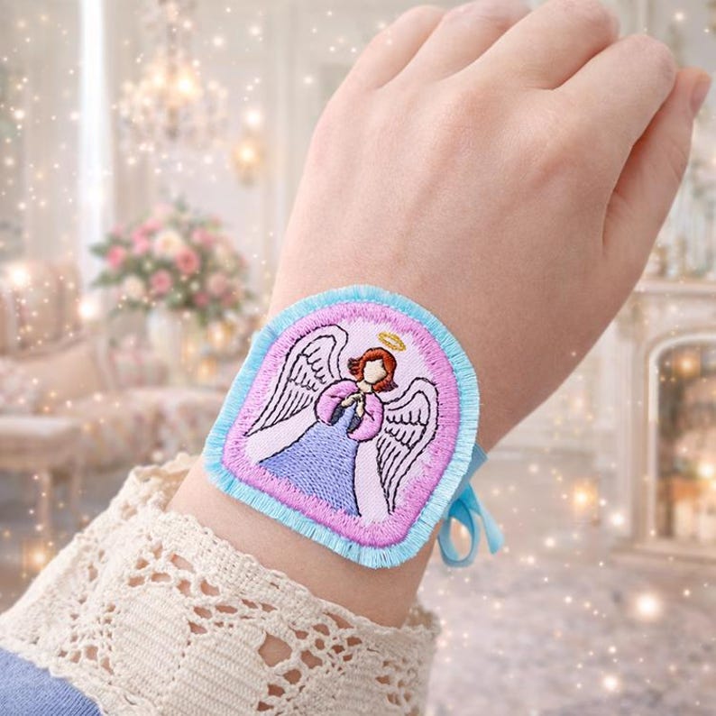Puede incluir: Brazalete bordado con un dise&ntilde;o de &aacute;ngel. El brazalete tiene un borde azul claro y un interior rosa con un &aacute;ngel con un vestido azul, halo dorado y detalles rosas. Se asegura con cintas azul claro.