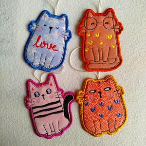 Può includere: Quattro ornamenti a forma di gatto colorati. Uno è blu con la parola "love", un altro è arancione con gli occhiali, un gatto rosa con strisce nere e un gatto arancione con cuori blu. Ognuno ha un cordino bianco per appenderlo.