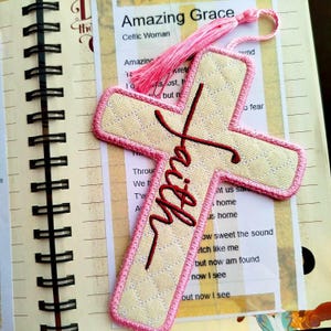 May include: A pink and cream fabric bookmark shaped like a cross with the word "Faith" embroidered in red. The bookmark has a pink tassel.