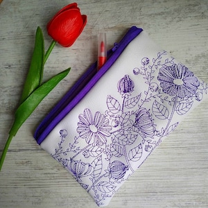 Peut inclure: Une pochette blanche à fermeture éclair avec une bordure violette et un motif floral brodé en violet. La pochette est posée sur une surface en bois blanc.