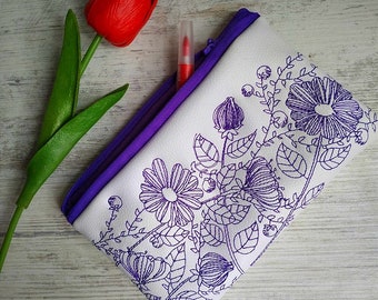 May include: A white zippered pouch with a purple trim and a floral design embroidered in purple. The pouch is lying on a white wooden surface.